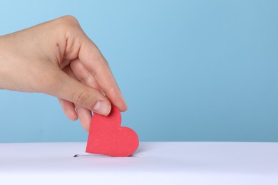 Woman putting red heart into slot of donation box against light blue background, closeup. Space for text