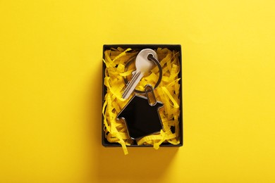 Key with trinket in shape of house and gift box on yellow background, top view. Housewarming party