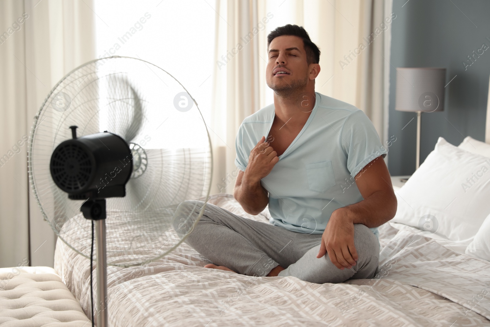 Photo of Man enjoying air flow from fan on bed in room. Summer heat