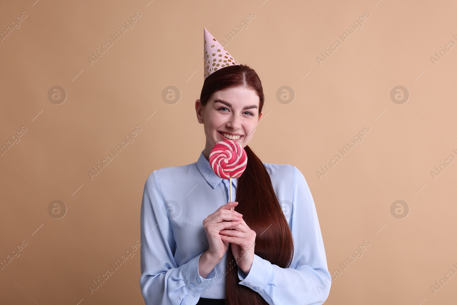 Photo of Happy woman in party hat with lollipop on beige background