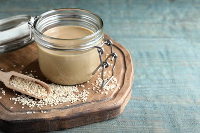 Photo of Jar of tasty sesame paste, seeds and scoop on light blue wooden table, space for text