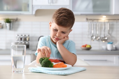 Photo of Unhappy little boy eating vegetables at table in kitchen