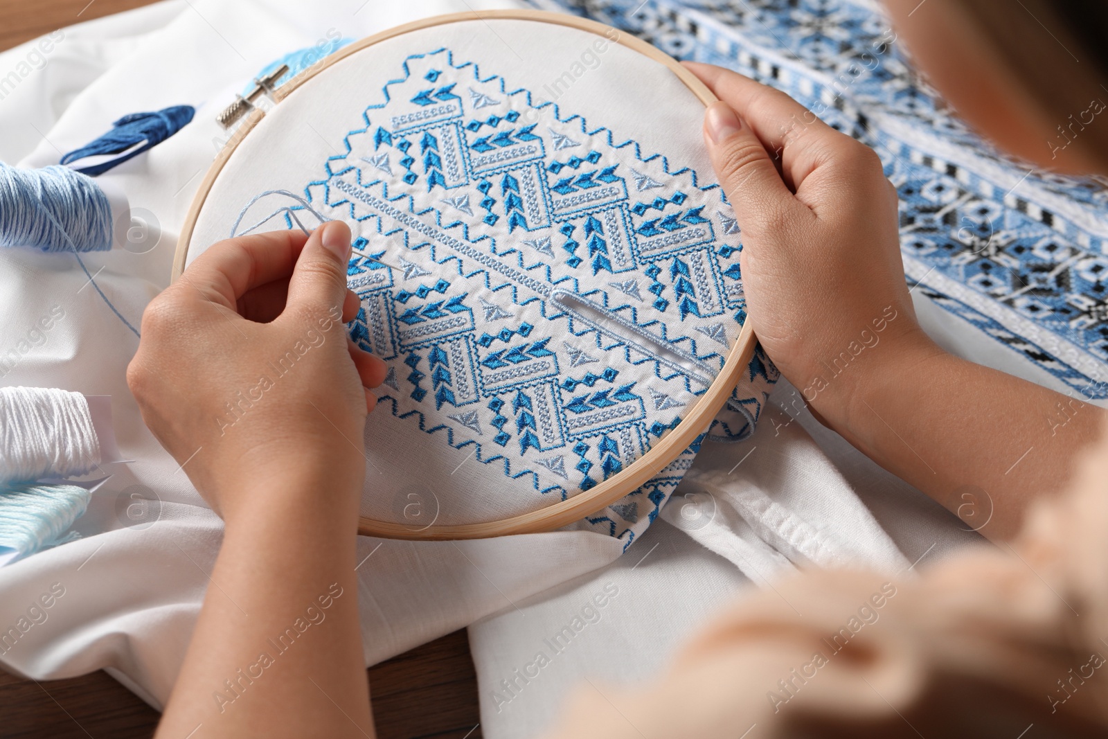 Photo of Woman embroidering white shirt with blue thread at wooden table, closeup. Ukrainian national clothes