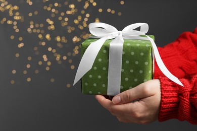 Photo of Christmas present. Woman holding gift box against grey background with blurred lights, closeup. Space for text
