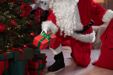 Photo of Santa Claus with gift and red bag near Christmas tree in room, closeup
