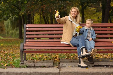 Happy mother and her son spending time together with dry leaves on wooden bench in autumn park