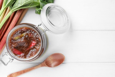 Photo of Jar of tasty rhubarb jam, stems and spoon on white wooden table, flat lay. Space for text