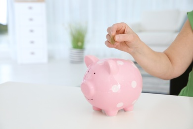 Photo of Mature woman putting money into piggy bank at table, closeup. Space for text