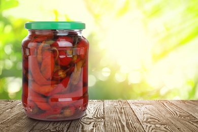 Jar of pickled red peppers on wooden table against blurred background, space for text