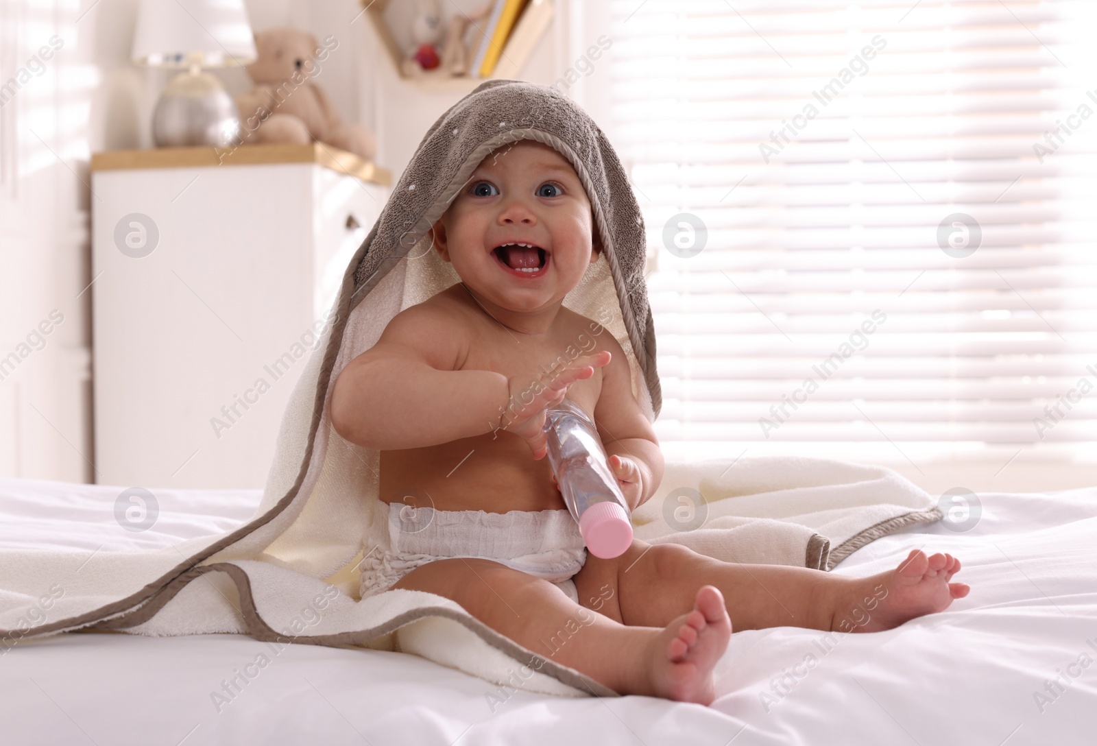 Photo of Cute little baby with towel and bottle of massage oil on bed at home