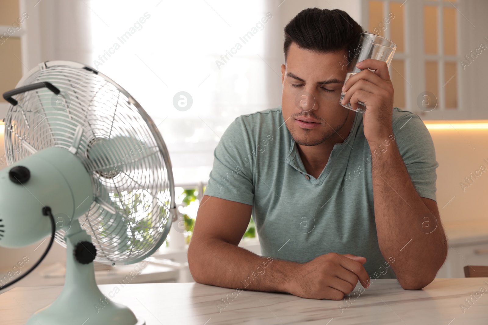 Photo of Man with glass of water and modern fan at table in kitchen. Summer heat