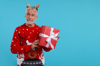 Photo of Senior man in Christmas sweater and reindeer headband holding gift on light blue background. Space for text
