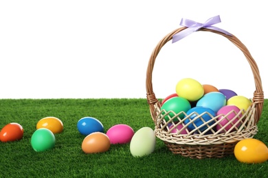 Photo of Wicker basket with bow and bright painted Easter eggs on green grass against white background