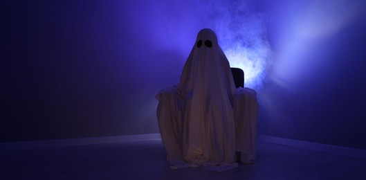 Photo of Creepy ghost. Woman covered with sheet sitting in armchair in blue light, space for text