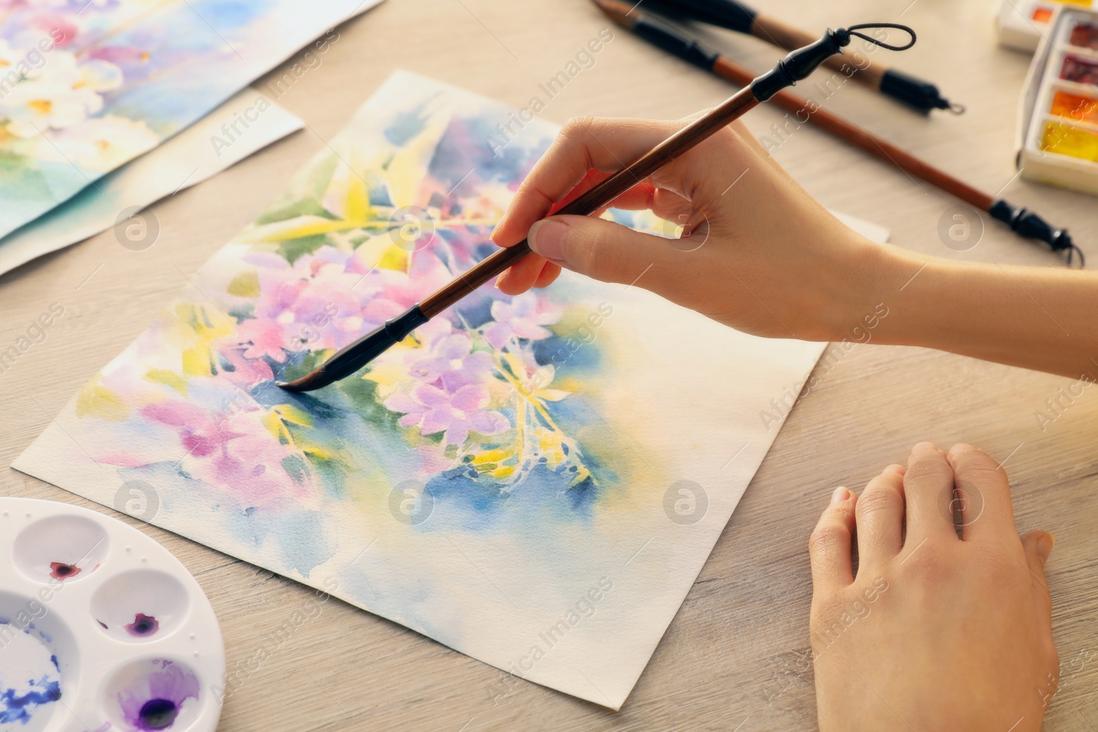 Photo of Woman painting flowers with watercolor at white wooden table, closeup. Creative artwork