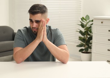 Photo of Sad man sitting at white table indoors. Space for text