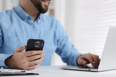 Photo of Young man using smartphone while working with laptop at white table in office, closeup