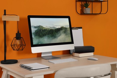 Photo of Modern computer, books, lamp and notebook on wooden desk near orange wall. Home office