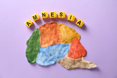 Photo of Word Amnesia made of yellow cubes and brain with sections on violet background, flat lay