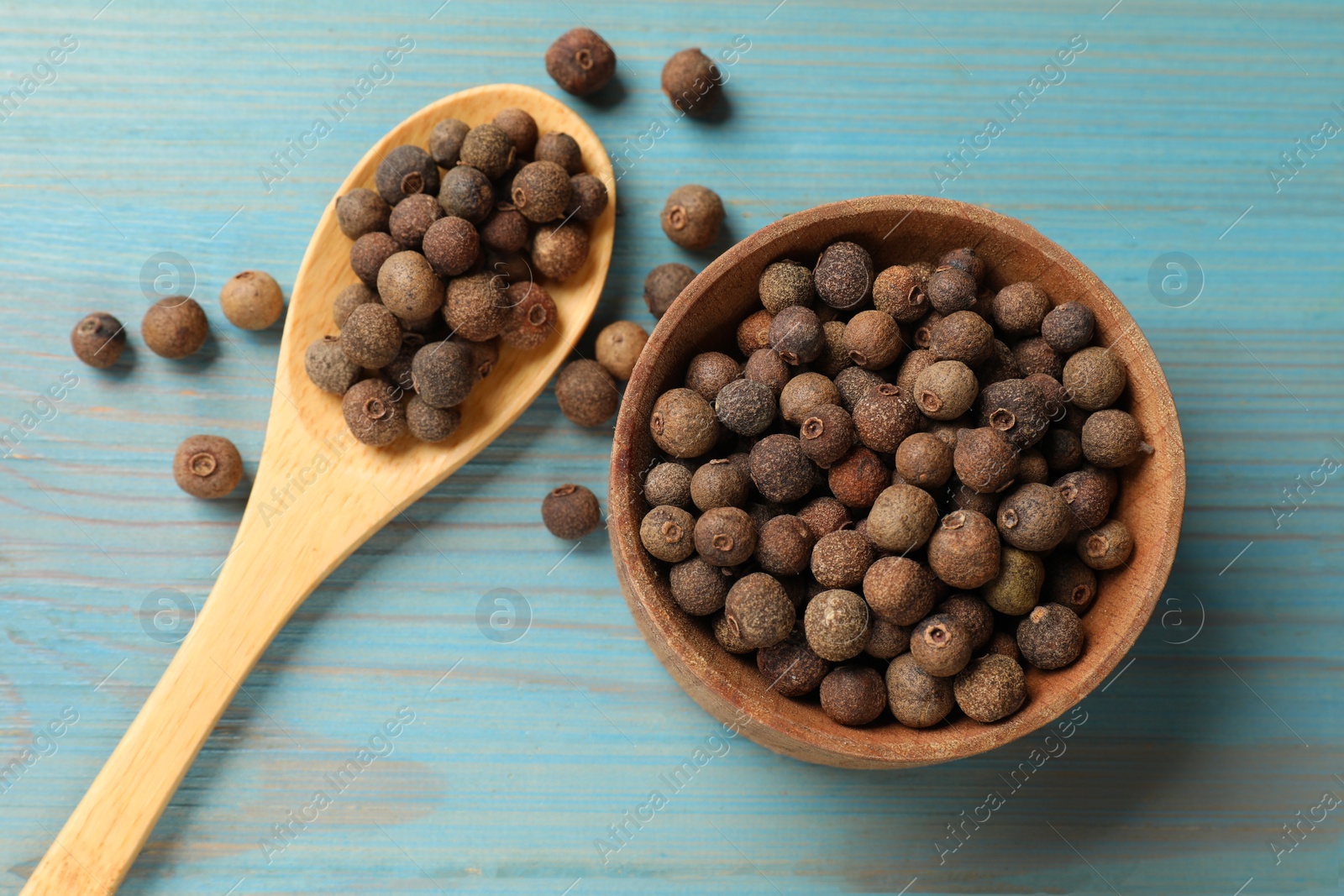 Photo of Aromatic allspice pepper grains in bowl and spoon on light blue wooden table, flat lay