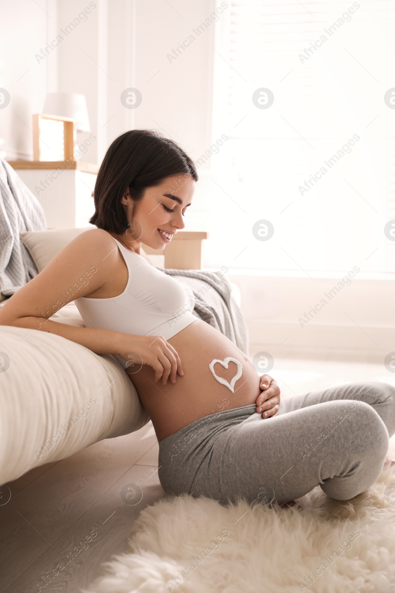 Photo of Young pregnant woman with cosmetic product on belly at home