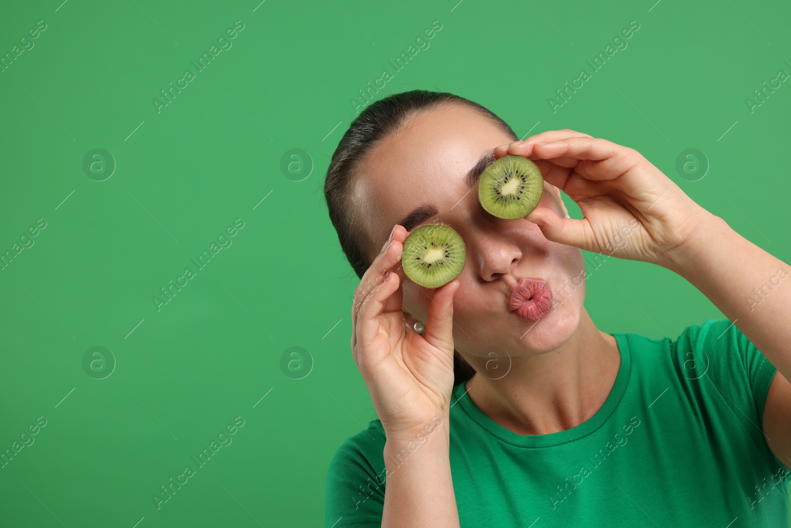Photo of Woman covering eyes with halves of kiwi on green background, space for text