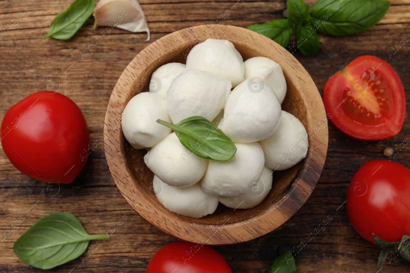 Photo of Delicious mozzarella balls in bowl, tomatoes and basil leaves on wooden table, flat lay.