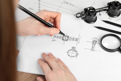 Jeweler drawing sketch of elegant ring on paper at wooden table, closeup