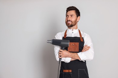 Smiling hairdresser wearing apron with dryer on light grey background, space for text