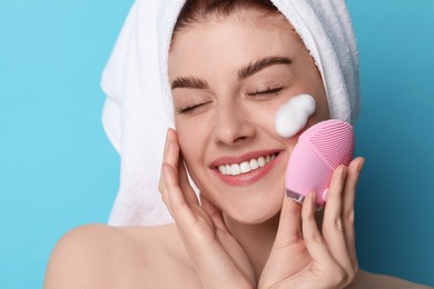 Photo of Young woman washing face with brush and cleansing foam on light blue background, closeup