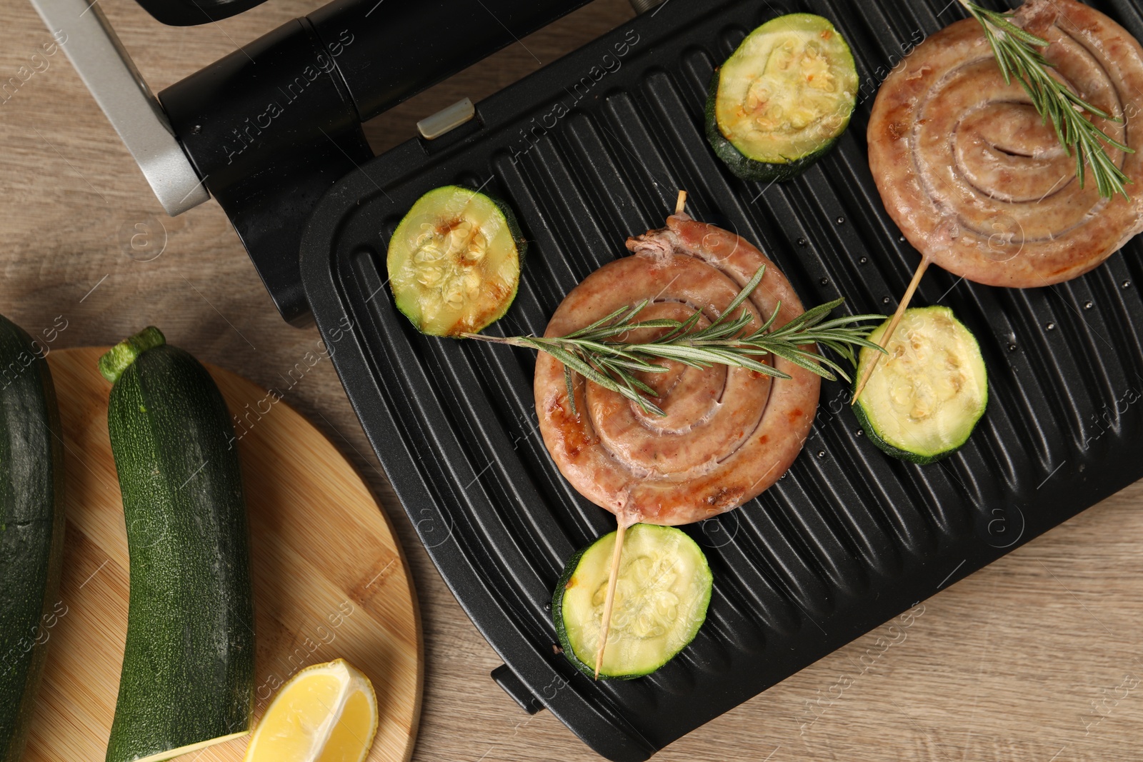 Photo of Electric grill with homemade sausages, zucchini and rosemary on wooden table, flat lay