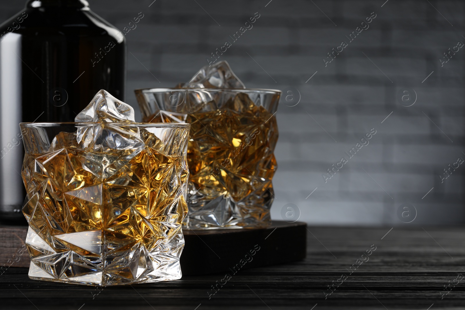 Photo of Whiskey with ice cubes in glasses and bottle on black wooden table, closeup. Space for text