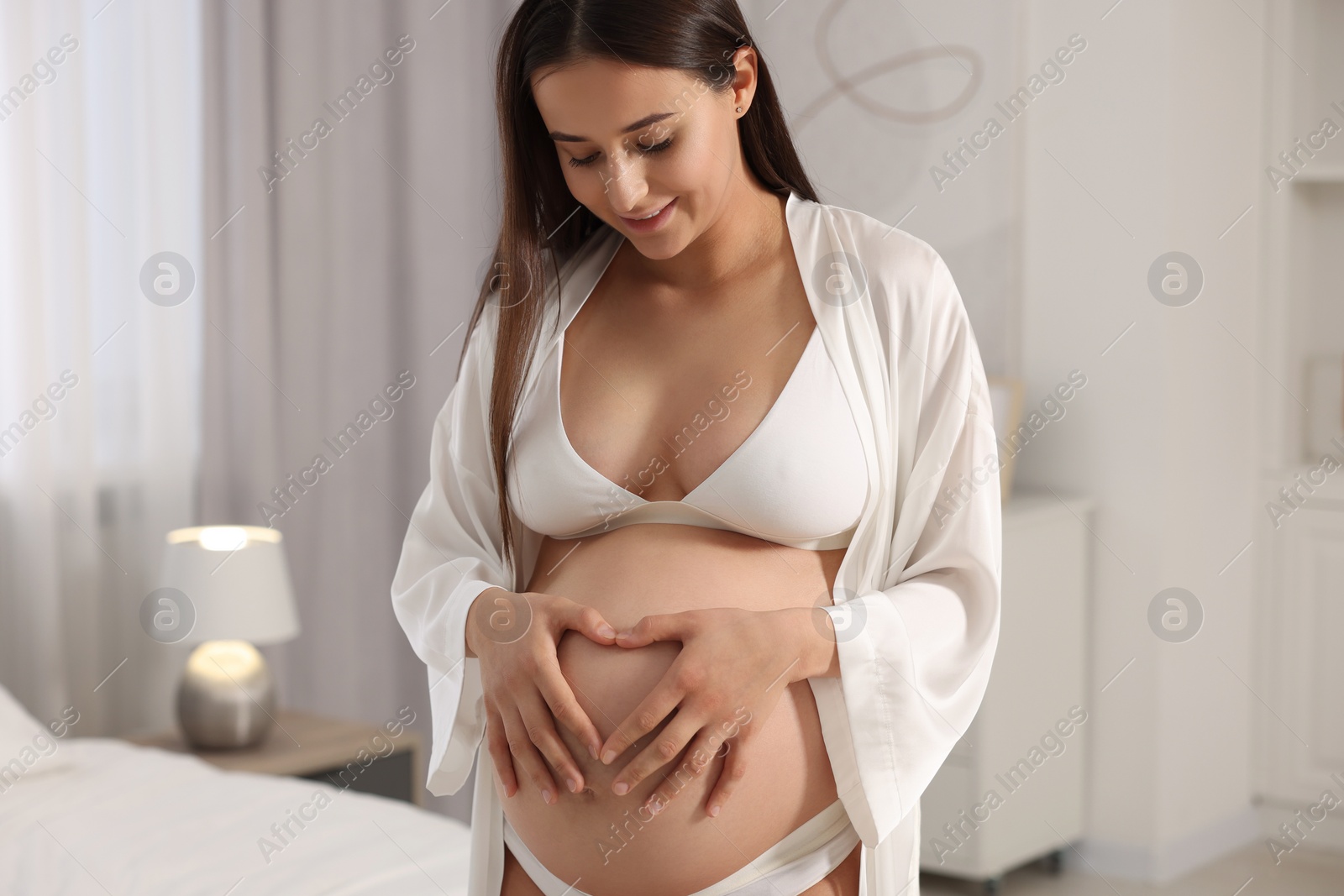 Photo of Beautiful pregnant woman in stylish comfortable underwear and robe making heart with hands on her belly in bedroom