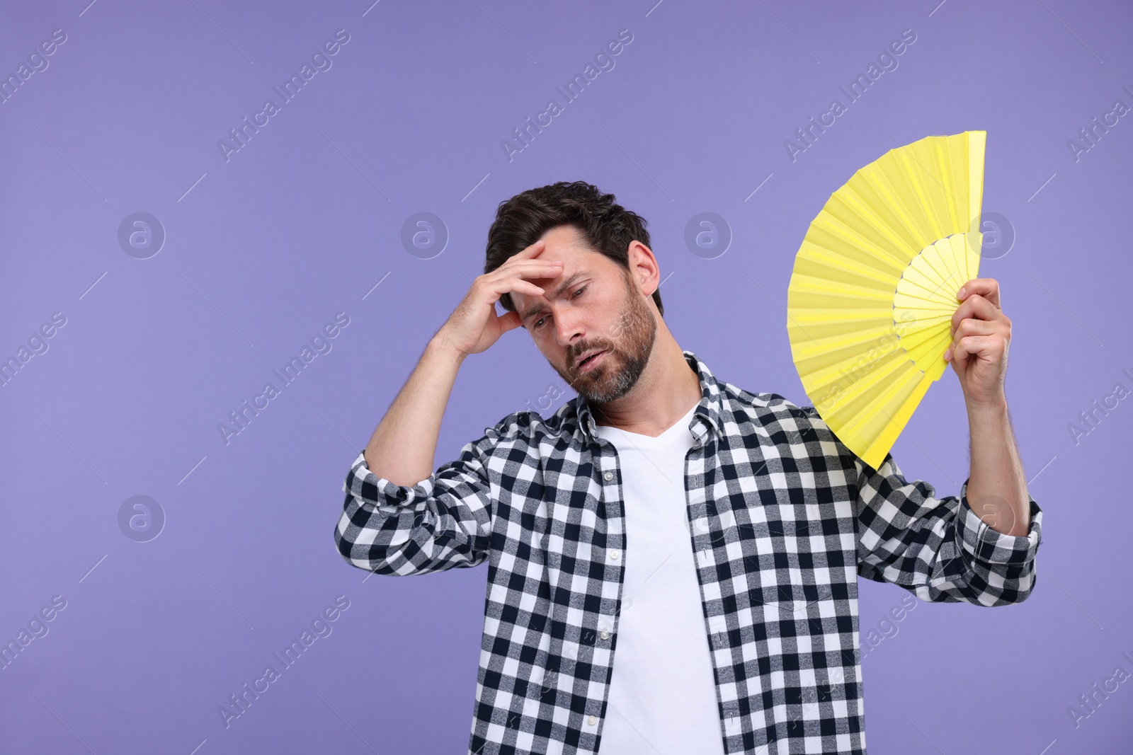 Photo of Unhappy man with hand fan suffering from heat on purple background. Space for text