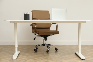 Photo of Stylish workplace with laptop and comfortable armchair near white wall indoors. Interior design