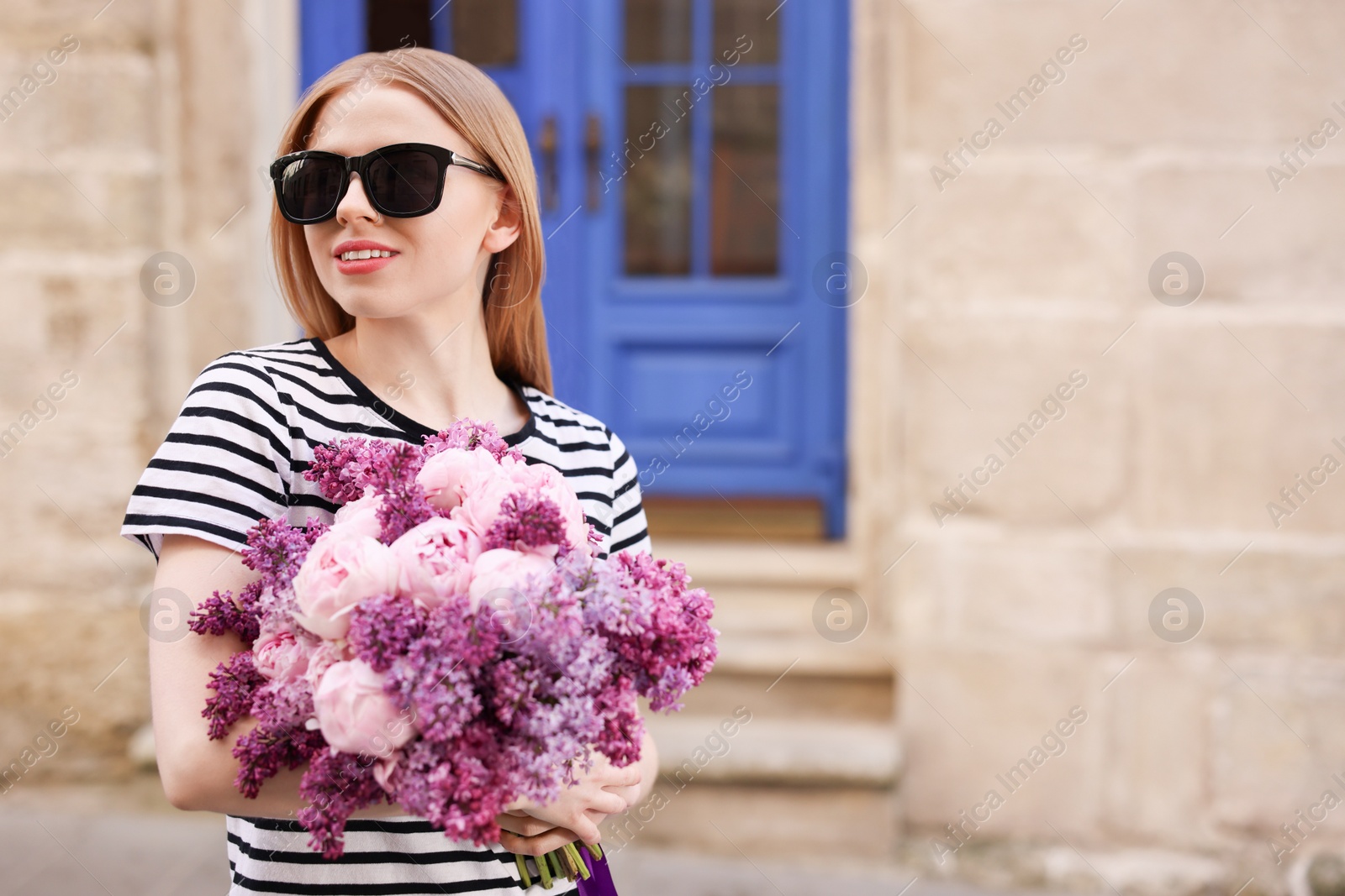 Photo of Beautiful woman in sunglasses with bouquet of spring flowers near building outdoors, space for text