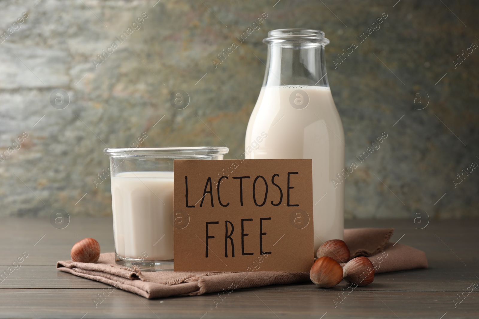 Photo of Milk in glassware, card with phrase Lactose free and hazelnuts on wooden table