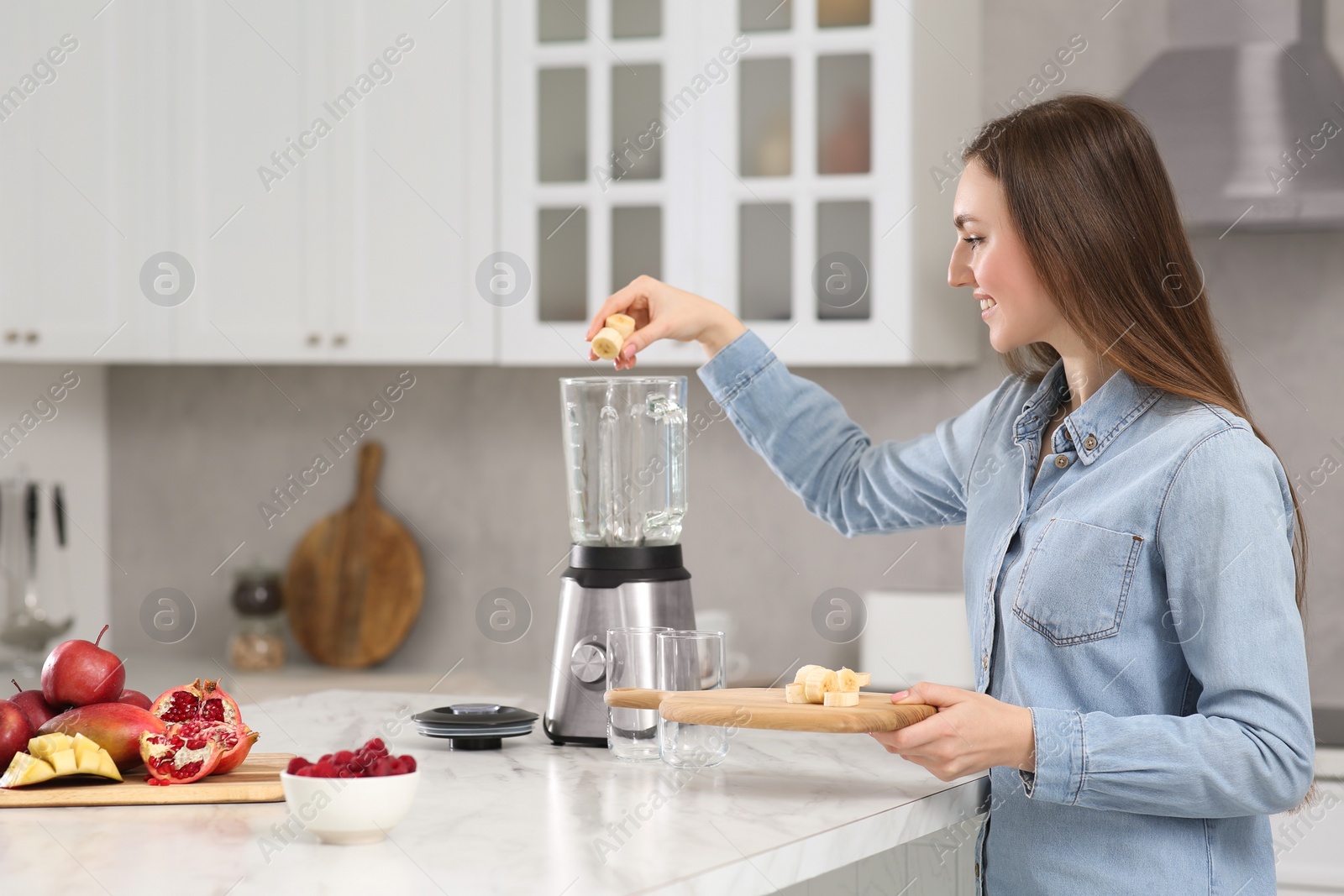 Photo of Beautiful young woman adding banana into blender for tasty smoothie in kitchen