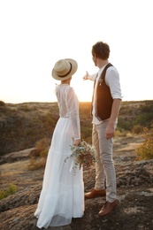 Happy newlyweds with beautiful field bouquet standing on rock at sunset