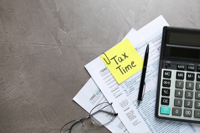 Photo of Reminder note with words TAX TIME, documents, glasses and calculator on grey table, flat lay. Space for text