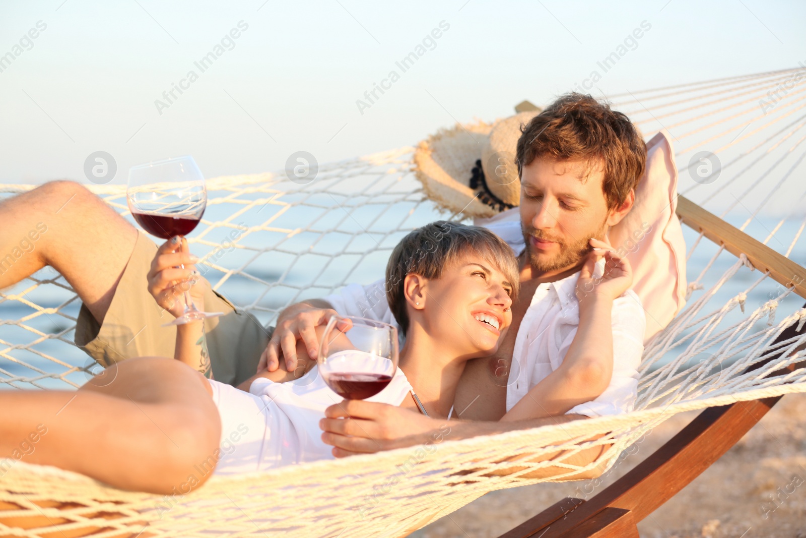Photo of Young couple resting with glasses of wine in hammock on beach