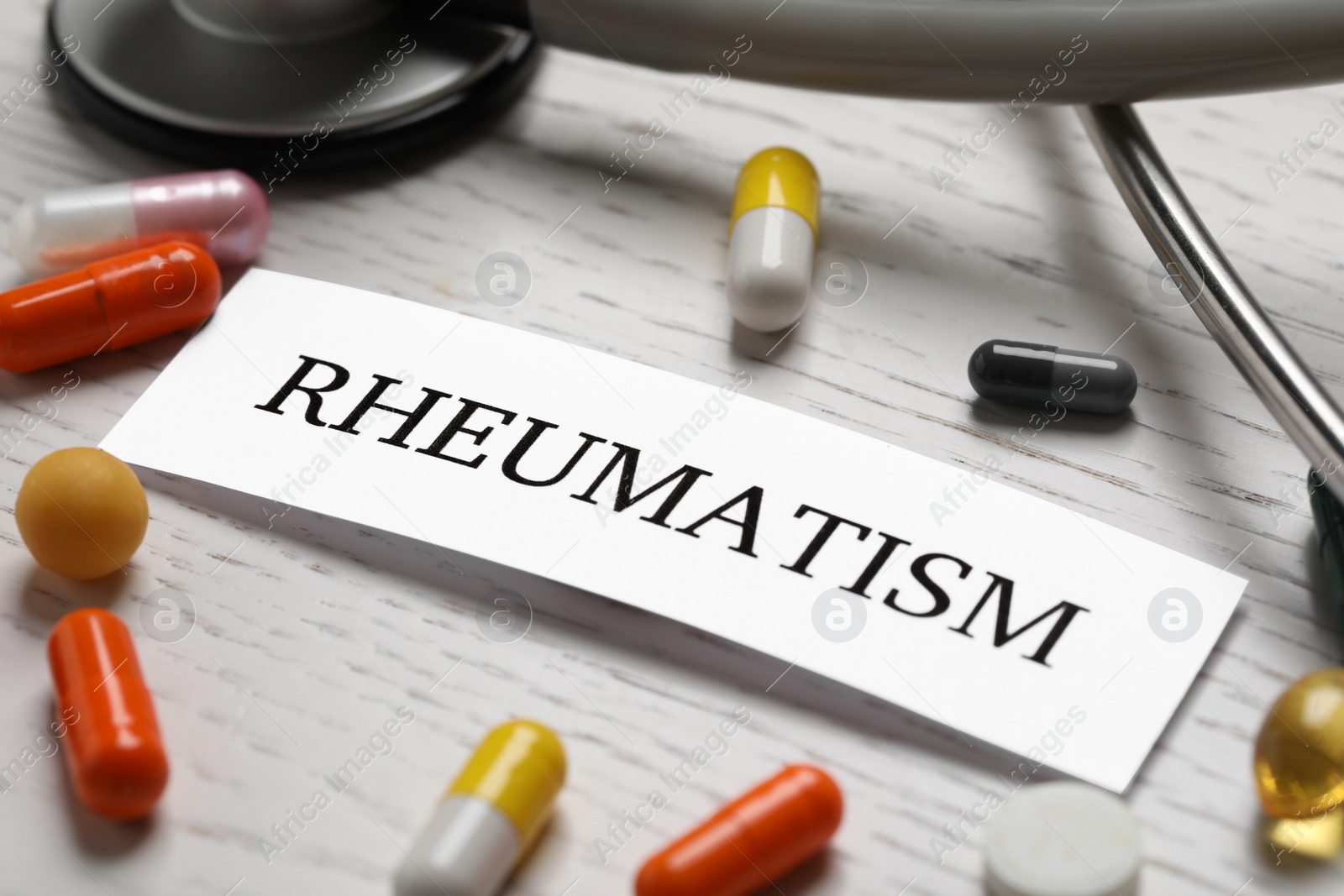 Photo of Sheet of paper with word Rheumatism and many different pills on white wooden table, closeup