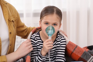 Photo of Mother covering her daughter with plaid while she using nebulizer for inhalation at home