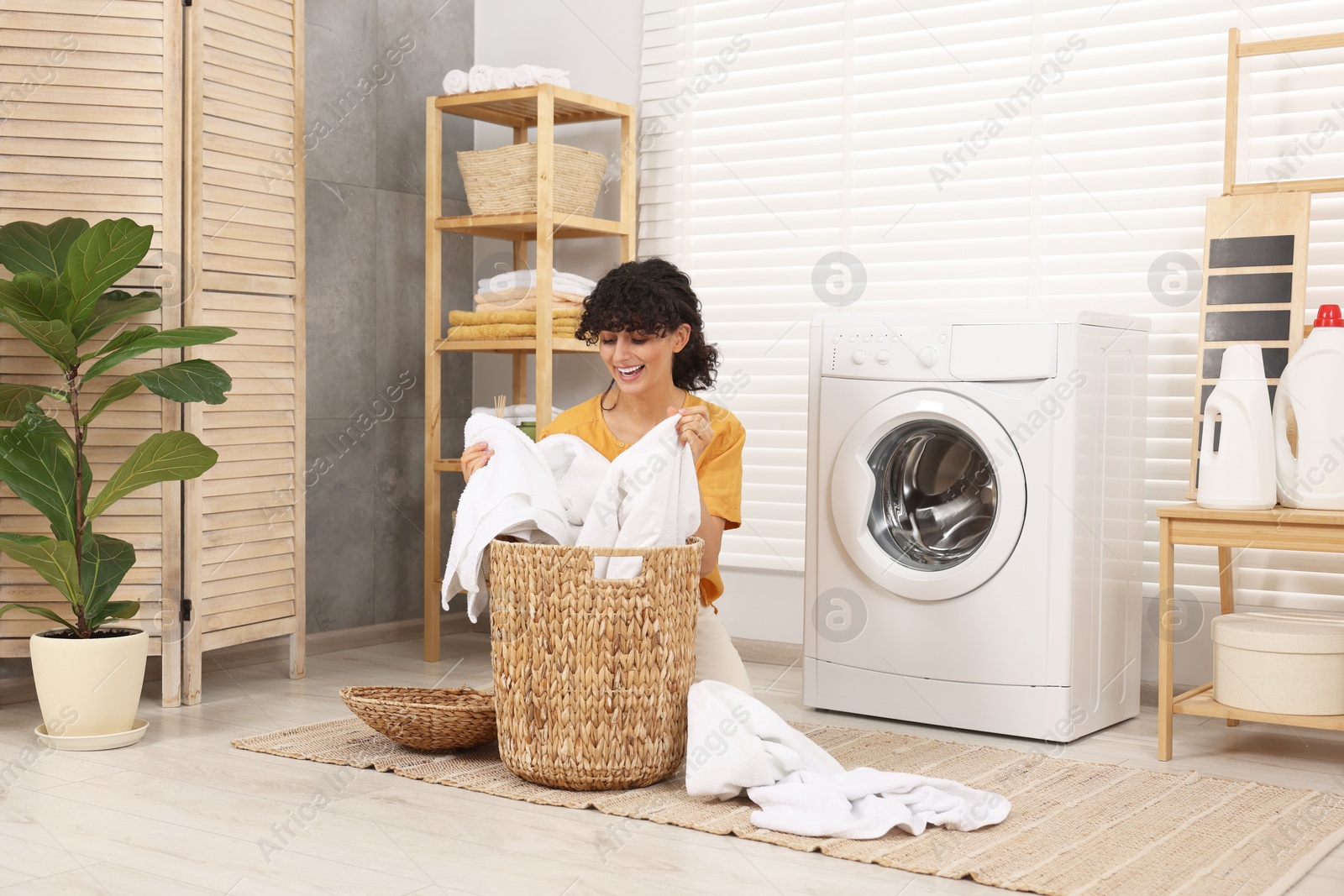 Photo of Happy woman taking laundry from basket near washing machine indoors
