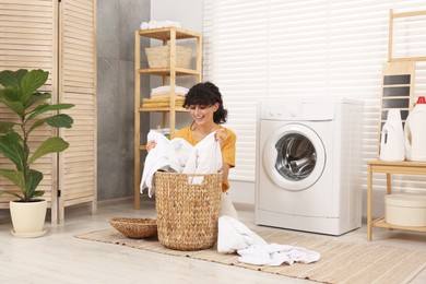 Happy woman taking laundry from basket near washing machine indoors