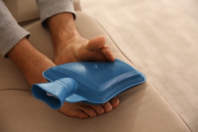 Photo of Man warming feet with hot water bottle on sofa, closeup