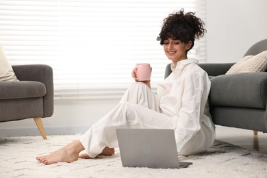 Beautiful young woman in stylish pyjama with cup of drink and laptop on floor at home