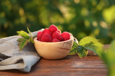 Photo of Tasty ripe raspberries in bowl and green leaves on wooden table outdoors