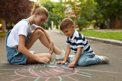 Photo of Teen nanny and cute little boy drawing sun with chalks on asphalt