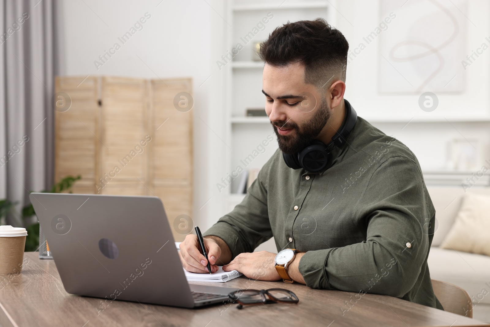 Photo of E-learning. Young man taking notes during online lesson at wooden table indoors